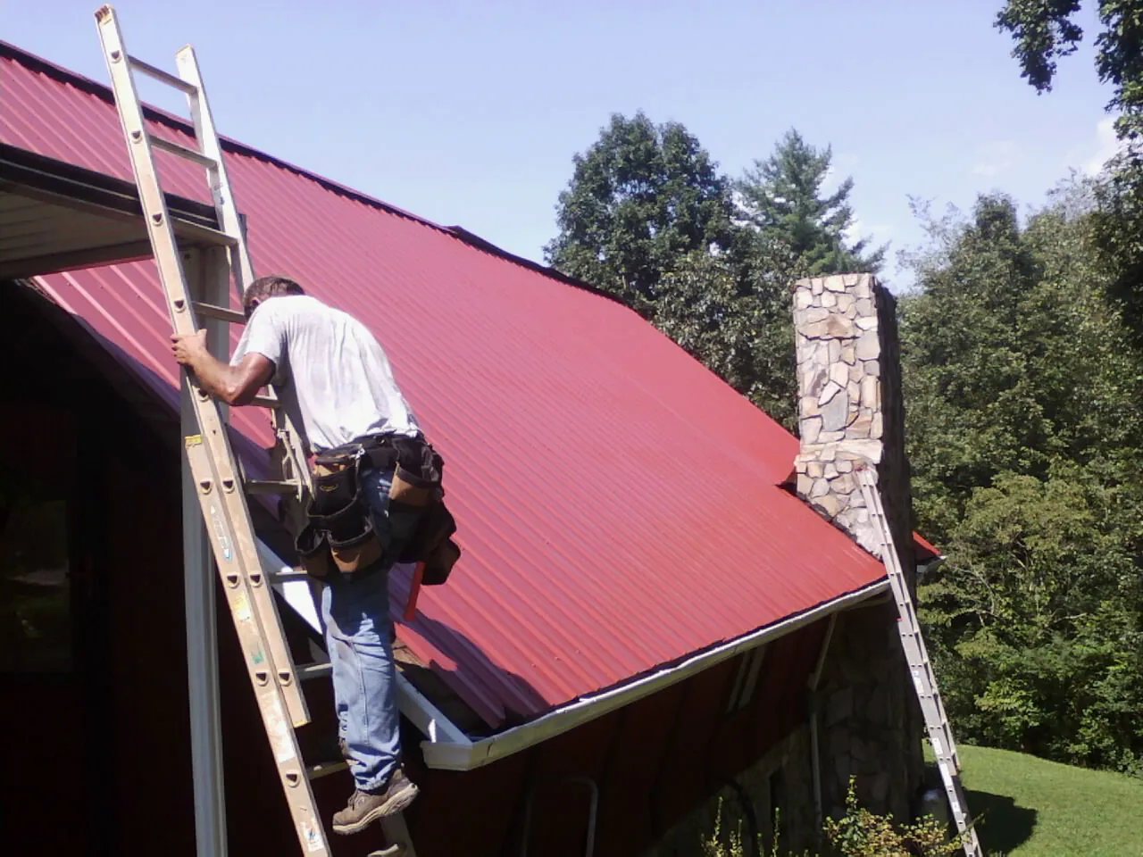 Roofer on a ladder beside a red metal roof and stone chimney — Soffit & Fascia Repair by Roofing Brothers in Cheney